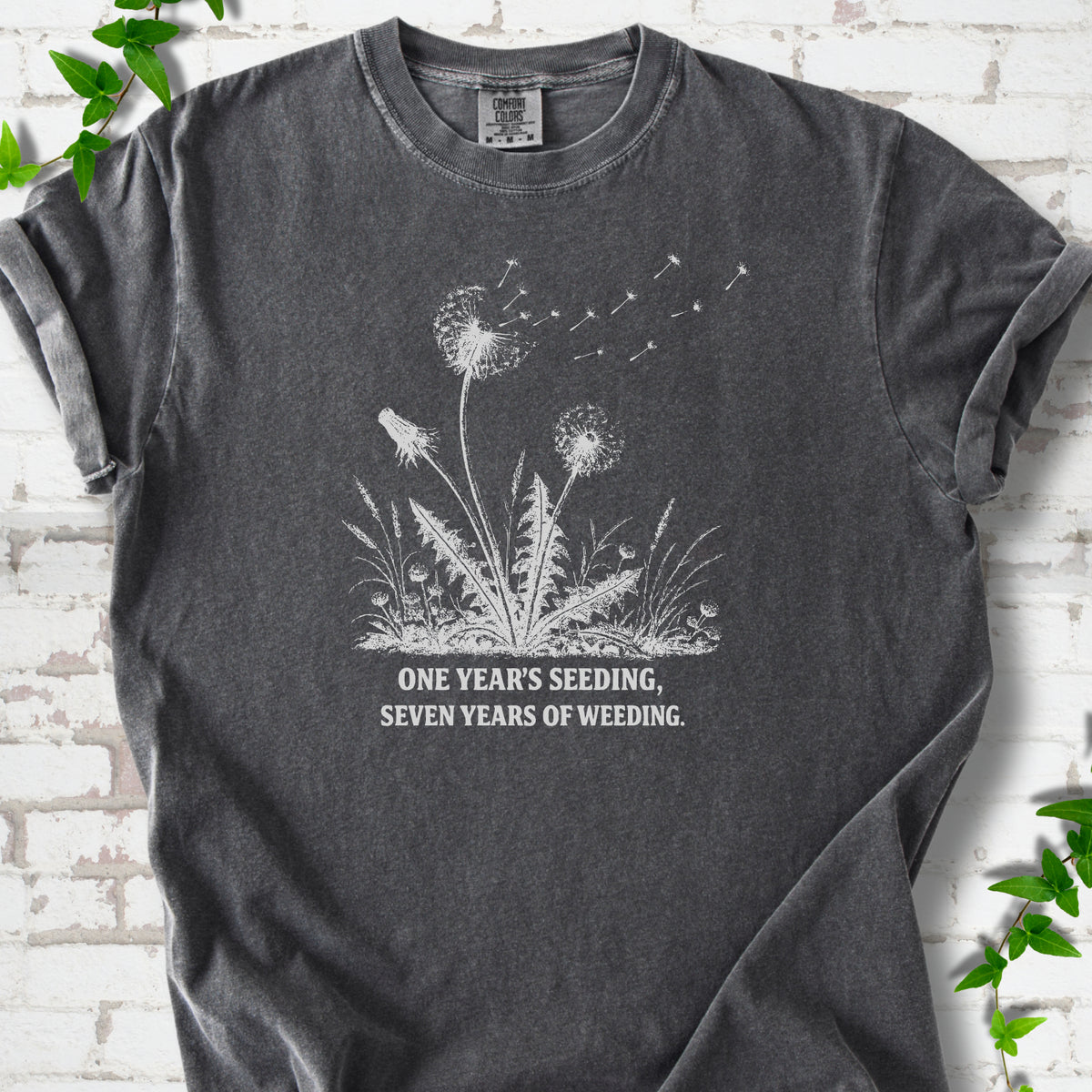 Pepper T-shirt flat lay with dandelion seed heads, drifting seeds and 'One year's seeding, seven years of weeding' lettering.