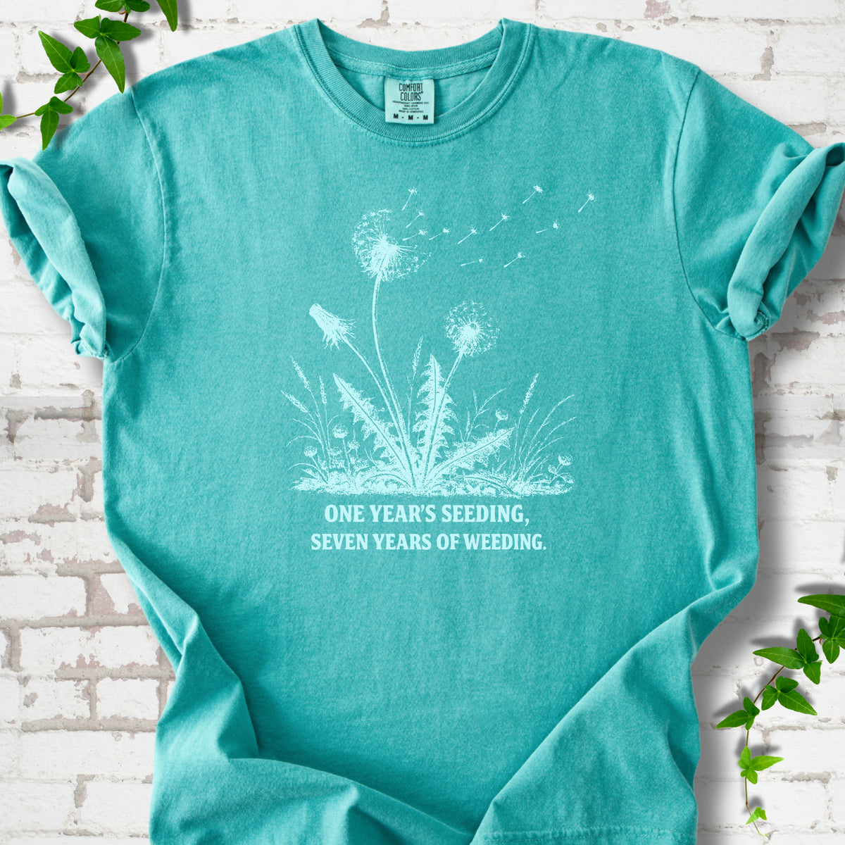 Seafoam T-shirt flat lay with dandelion seed heads, drifting seeds and 'One year's seeding, seven years of weeding' lettering.