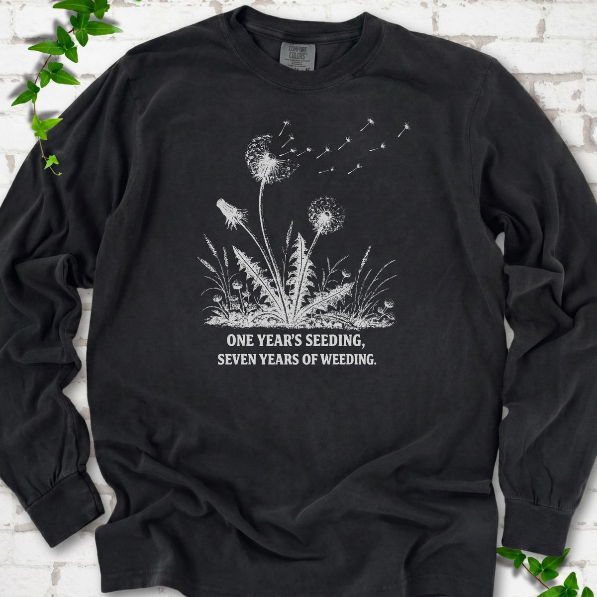 Black long sleeve T-shirt flat lay with dandelion seed heads, drifting seeds and 'One year's seeding, seven years of weeding' lettering.