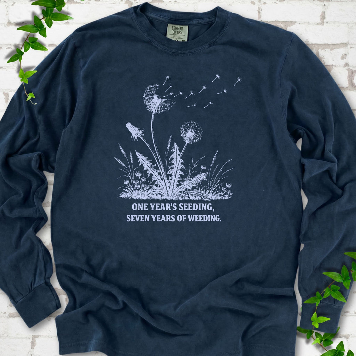 Midnight long sleeve T-shirt flat lay with dandelion seed heads, drifting seeds and 'One year's seeding, seven years of weeding' lettering.
