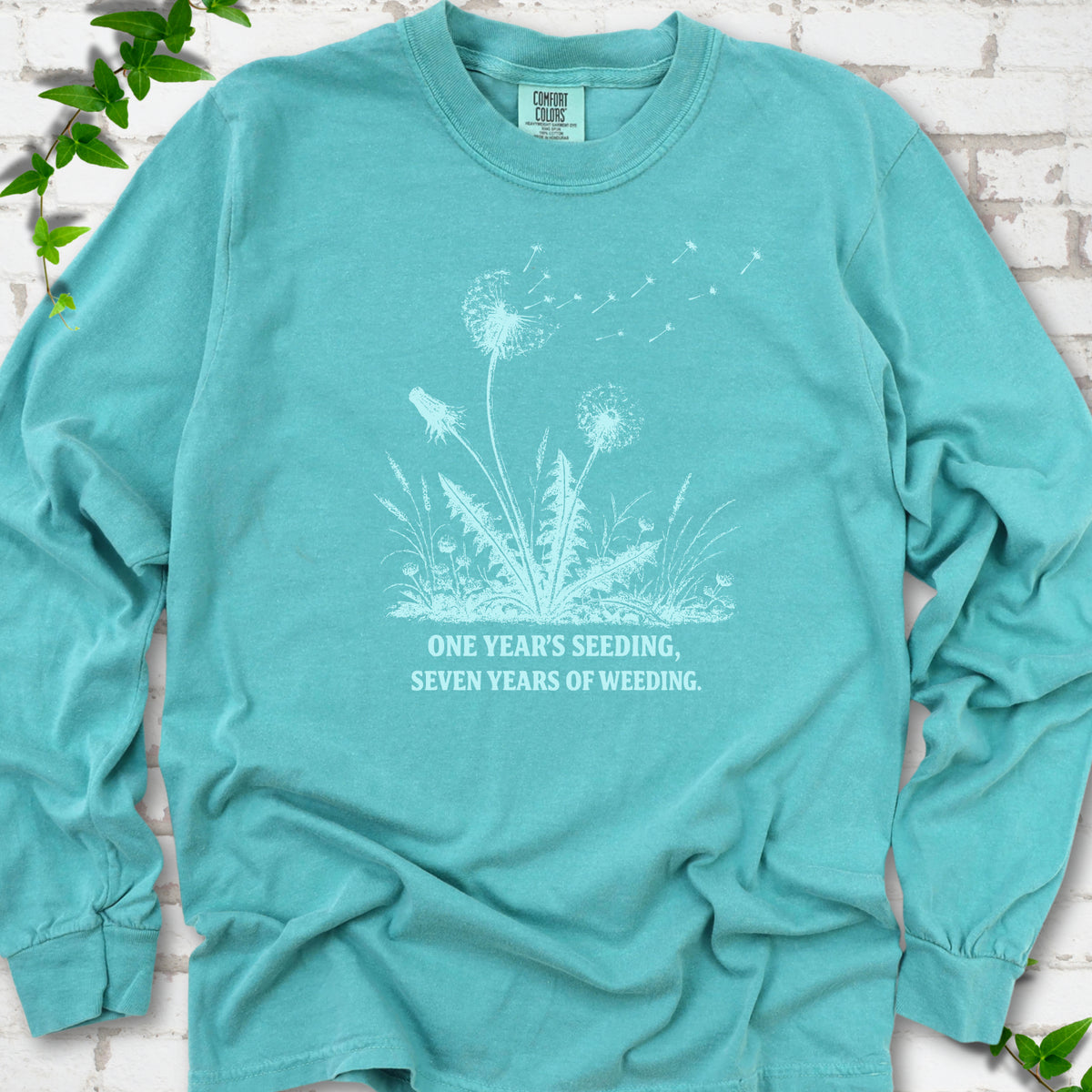 Seafoam long sleeve T-shirt flat lay with dandelion seed heads, drifting seeds and 'One year's seeding, seven years of weeding' lettering.