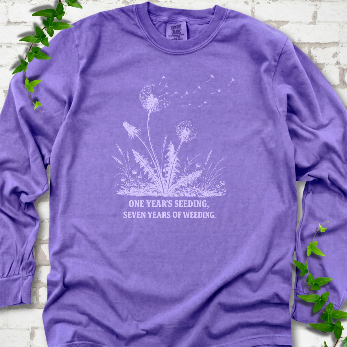 Violet long sleeve T-shirt flat lay with dandelion seed heads, drifting seeds and 'One year's seeding, seven years of weeding' lettering.