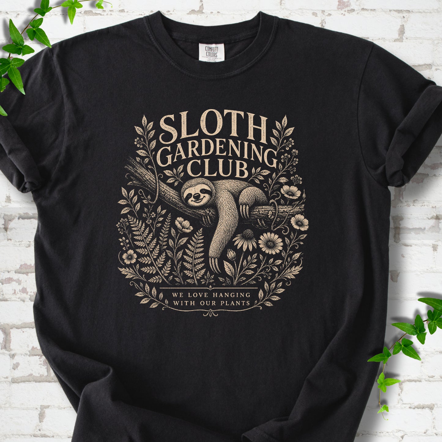 Black Comfort Colors® T-shirt front mockup featuring a detailed cream-colored engraving-style illustration of a smiling sloth resting on a tree branch, surrounded by wild flowers and ferns.
