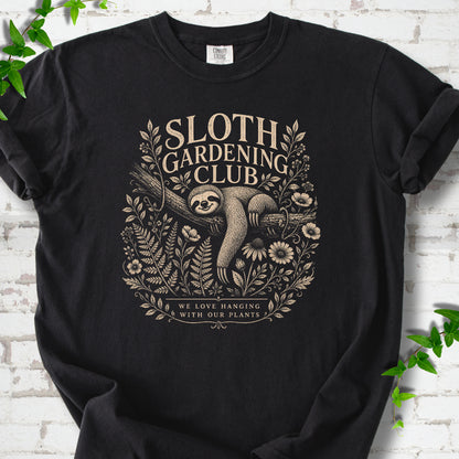 Black Comfort Colors® T-shirt front mockup featuring a detailed cream-colored engraving-style illustration of a smiling sloth resting on a tree branch, surrounded by wild flowers and ferns.