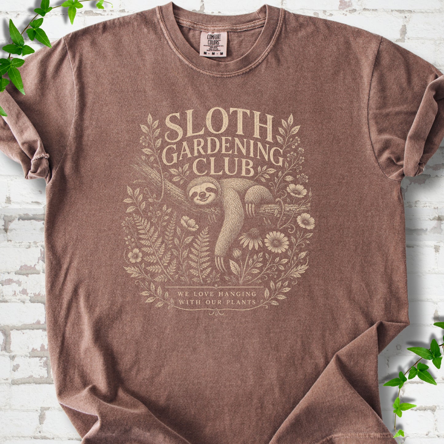 Espresso Comfort Colors® T-shirt front mockup featuring a detailed cream-colored engraving-style illustration of a smiling sloth resting on a tree branch, surrounded by wild flowers and ferns.