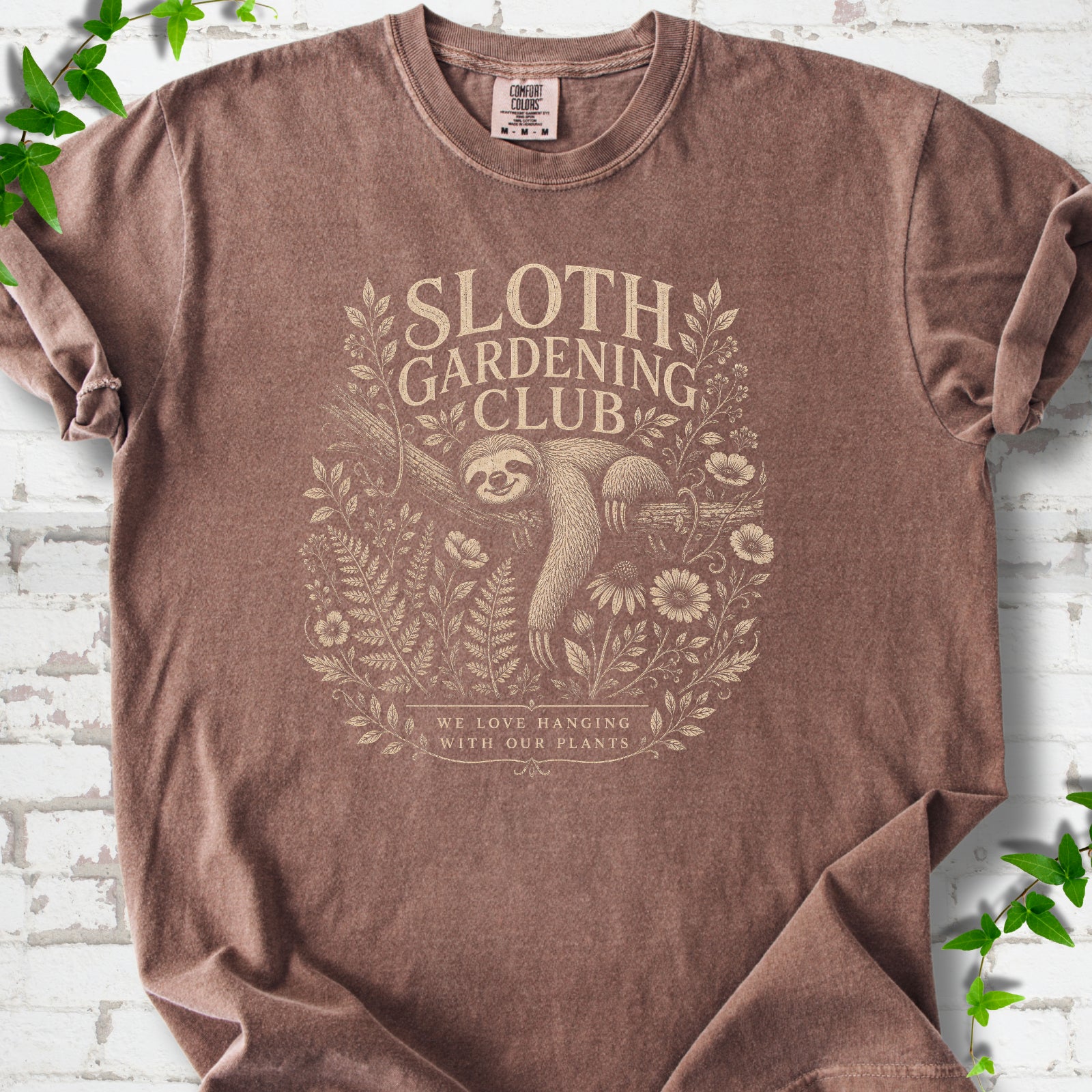 Espresso Comfort Colors® T-shirt front mockup featuring a detailed cream-colored engraving-style illustration of a smiling sloth resting on a tree branch, surrounded by wild flowers and ferns.