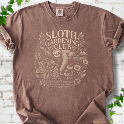 Espresso Comfort Colors® T-shirt front mockup featuring a detailed cream-colored engraving-style illustration of a smiling sloth resting on a tree branch, surrounded by wild flowers and ferns.