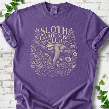 Grape Comfort Colors® T-shirt front mockup featuring a detailed cream-colored engraving-style illustration of a smiling sloth resting on a tree branch, surrounded by wild flowers and ferns.