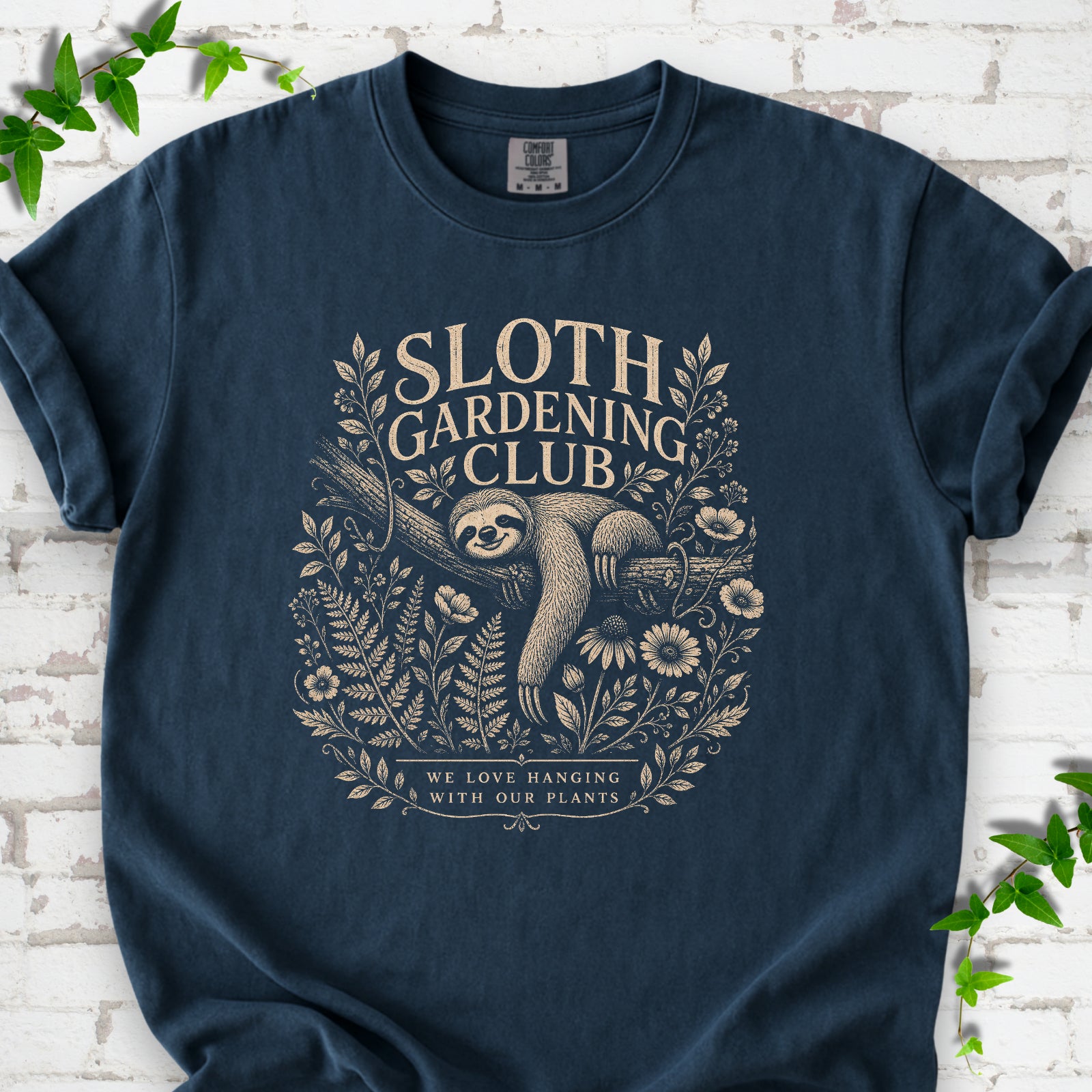 Midnight Comfort Colors® T-shirt front mockup featuring a detailed cream-colored engraving-style illustration of a smiling sloth resting on a tree branch, surrounded by wild flowers and ferns.