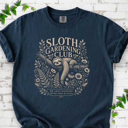 Midnight Comfort Colors® T-shirt front mockup featuring a detailed cream-colored engraving-style illustration of a smiling sloth resting on a tree branch, surrounded by wild flowers and ferns.