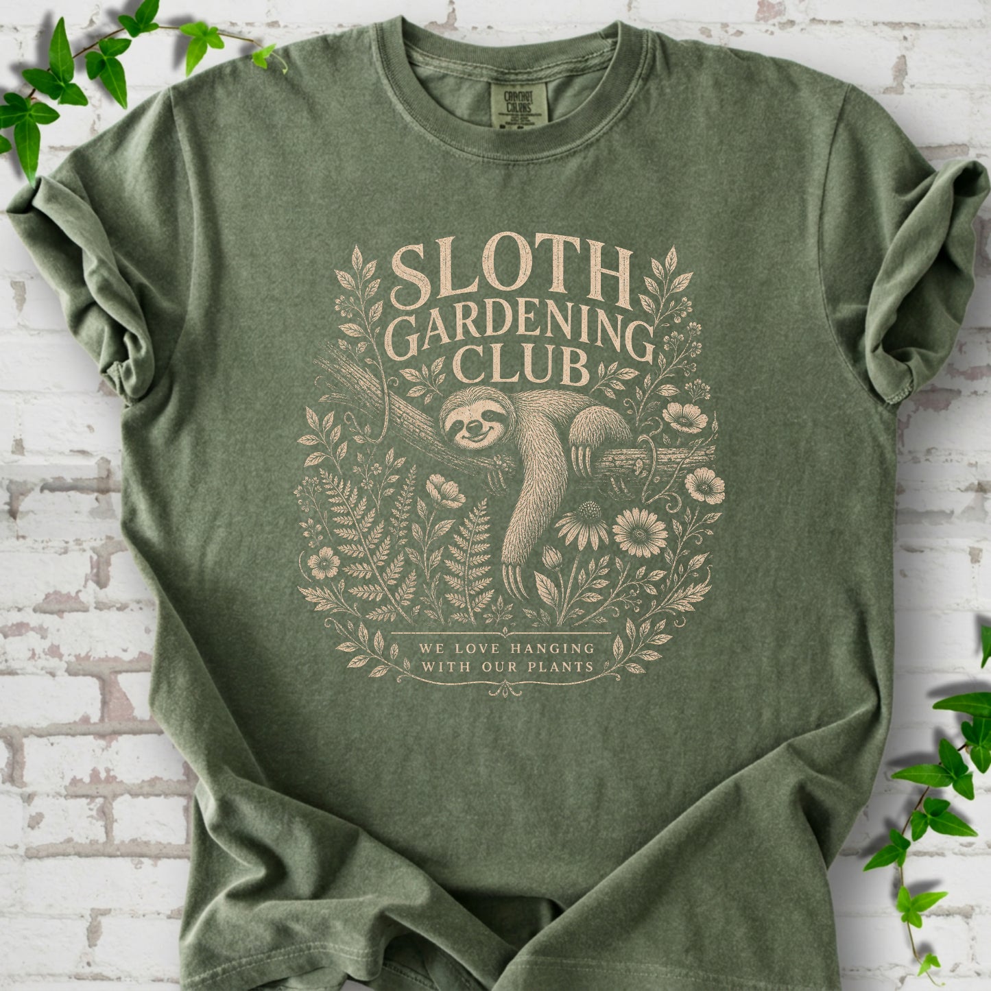 Moss Comfort Colors® T-shirt front mockup featuring a detailed cream-colored engraving-style illustration of a smiling sloth resting on a tree branch, surrounded by wild flowers and ferns.