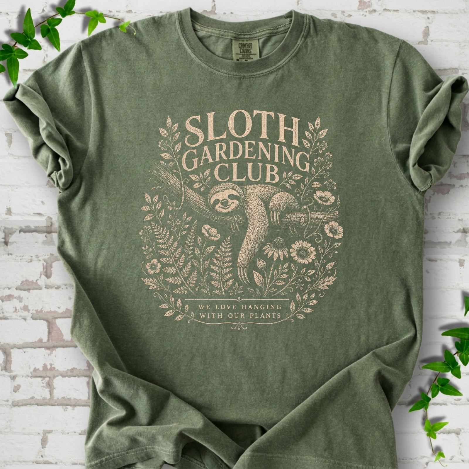 Moss Comfort Colors® T-shirt front mockup featuring a detailed cream-colored engraving-style illustration of a smiling sloth resting on a tree branch, surrounded by wild flowers and ferns.
