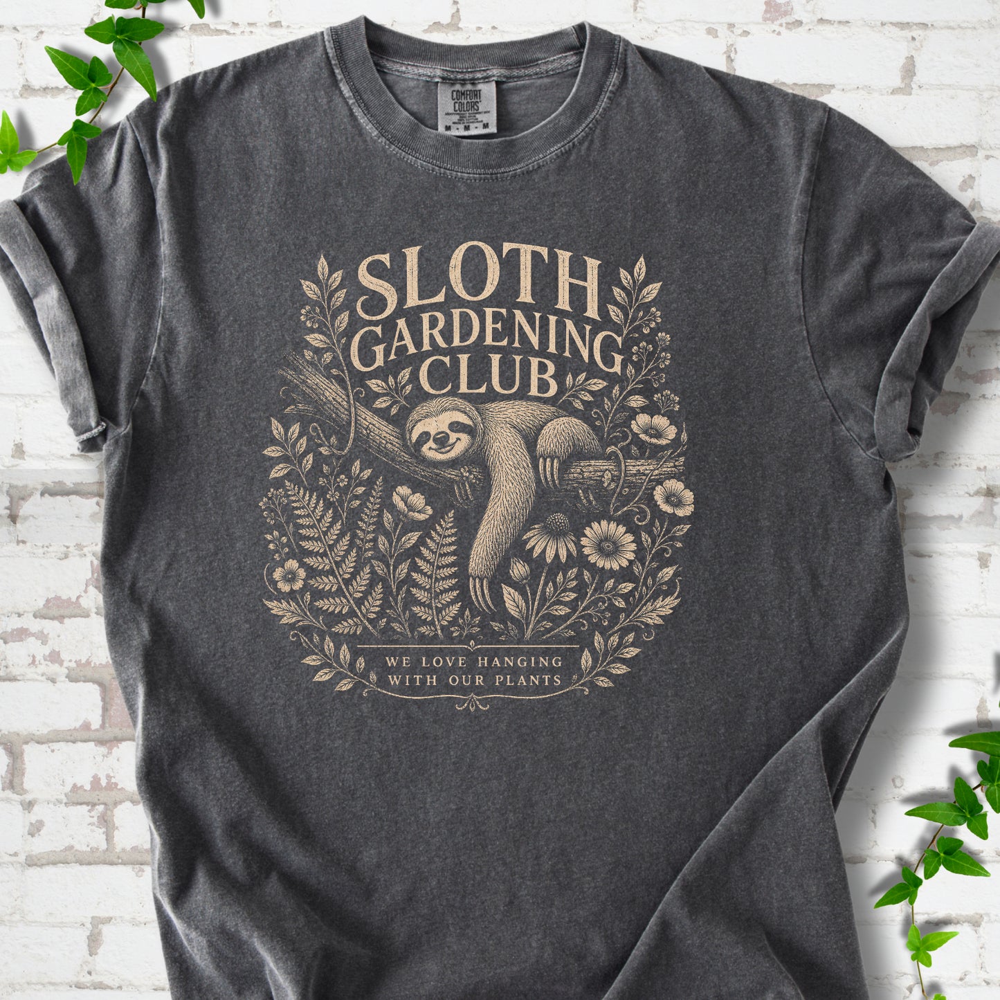 Pepper Comfort Colors® T-shirt front mockup featuring a detailed cream-colored engraving-style illustration of a smiling sloth resting on a tree branch, surrounded by wild flowers and ferns.