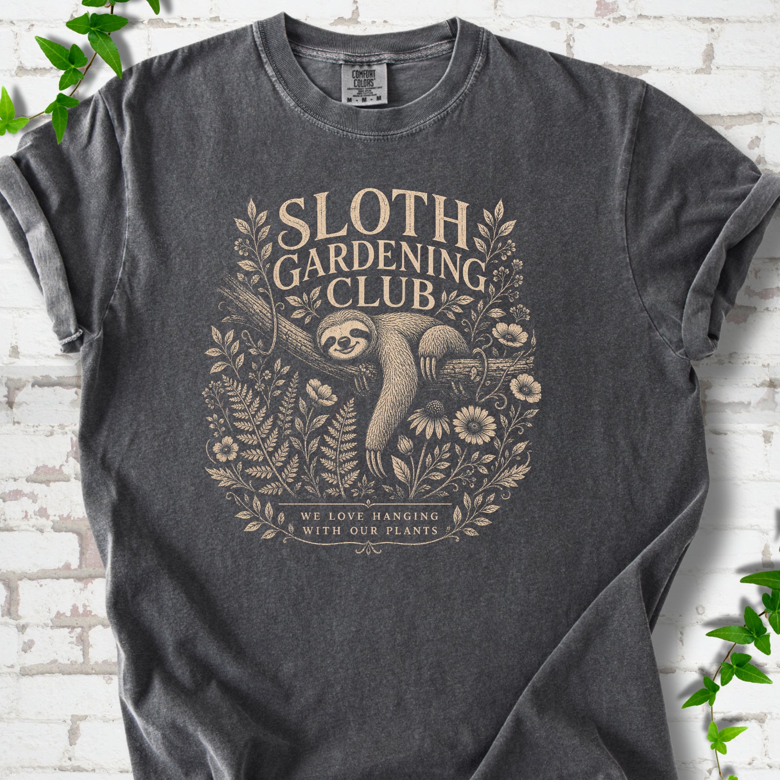 Pepper Comfort Colors® T-shirt front mockup featuring a detailed cream-colored engraving-style illustration of a smiling sloth resting on a tree branch, surrounded by wild flowers and ferns.