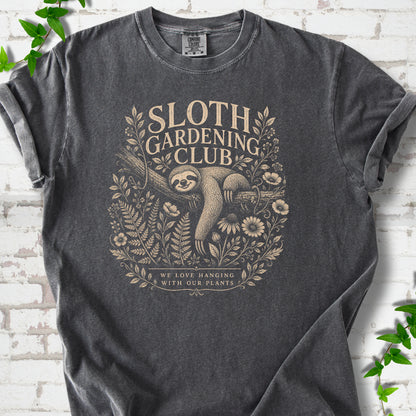 Pepper Comfort Colors® T-shirt front mockup featuring a detailed cream-colored engraving-style illustration of a smiling sloth resting on a tree branch, surrounded by wild flowers and ferns.