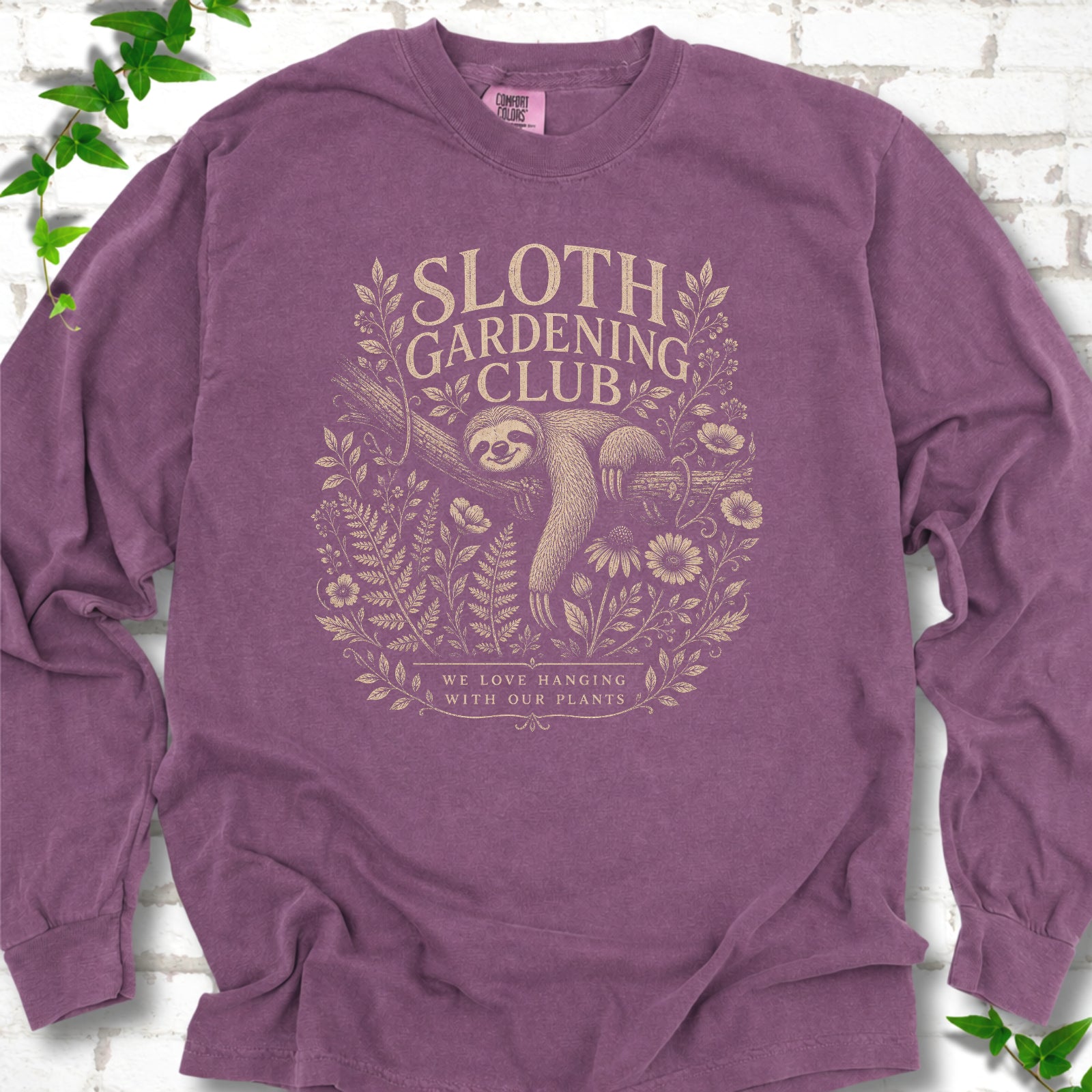 Berry Comfort Colors® long sleeve T-shirt front mockup featuring a detailed cream-colored engraving-style illustration of a smiling sloth resting on a tree branch, surrounded by wild flowers and ferns.