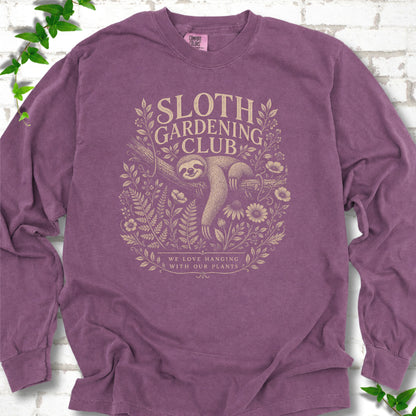 Berry Comfort Colors® long sleeve T-shirt front mockup featuring a detailed cream-colored engraving-style illustration of a smiling sloth resting on a tree branch, surrounded by wild flowers and ferns.