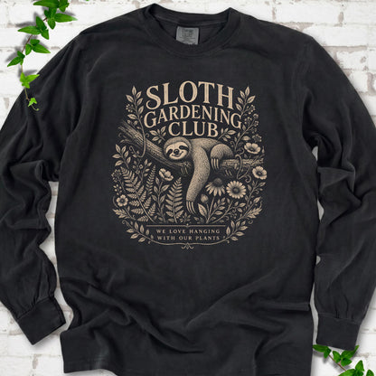 Black Comfort Colors® long sleeve T-shirt front mockup featuring a detailed cream-colored engraving-style illustration of a smiling sloth resting on a tree branch, surrounded by wild flowers and ferns.
