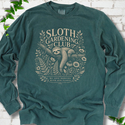 Blue Spruce Comfort Colors® long sleeve T-shirt front mockup featuring a detailed cream-colored engraving-style illustration of a smiling sloth resting on a tree branch, surrounded by wild flowers and ferns.
