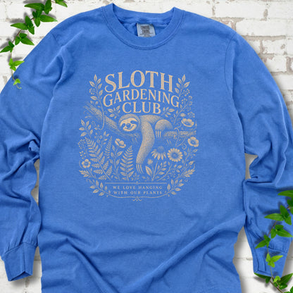 Flo Blue Comfort Colors® long sleeve T-shirt front mockup featuring a detailed cream-colored engraving-style illustration of a smiling sloth resting on a tree branch, surrounded by wild flowers and ferns.
