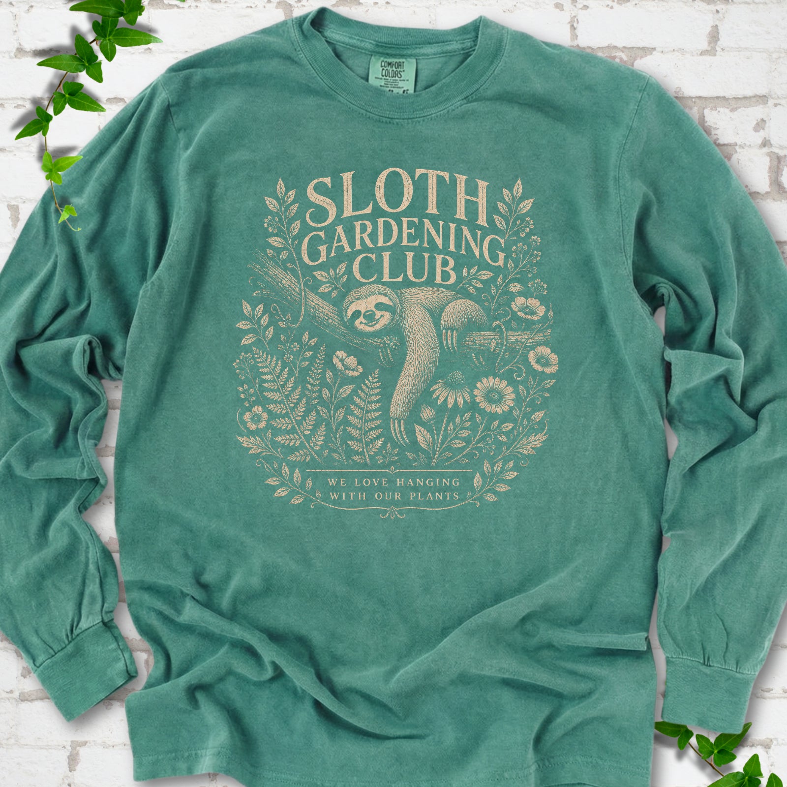 Light Green Comfort Colors® long sleeve T-shirt front mockup featuring a detailed cream-colored engraving-style illustration of a smiling sloth resting on a tree branch, surrounded by wild flowers and ferns.