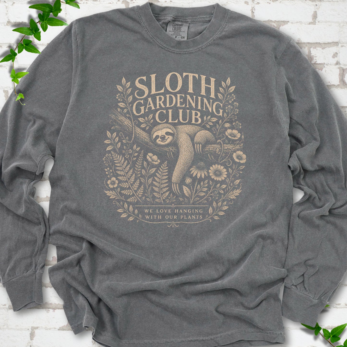 Pepper Comfort Colors® long sleeve T-shirt front mockup featuring a detailed cream-colored engraving-style illustration of a smiling sloth resting on a tree branch, surrounded by wild flowers and ferns.