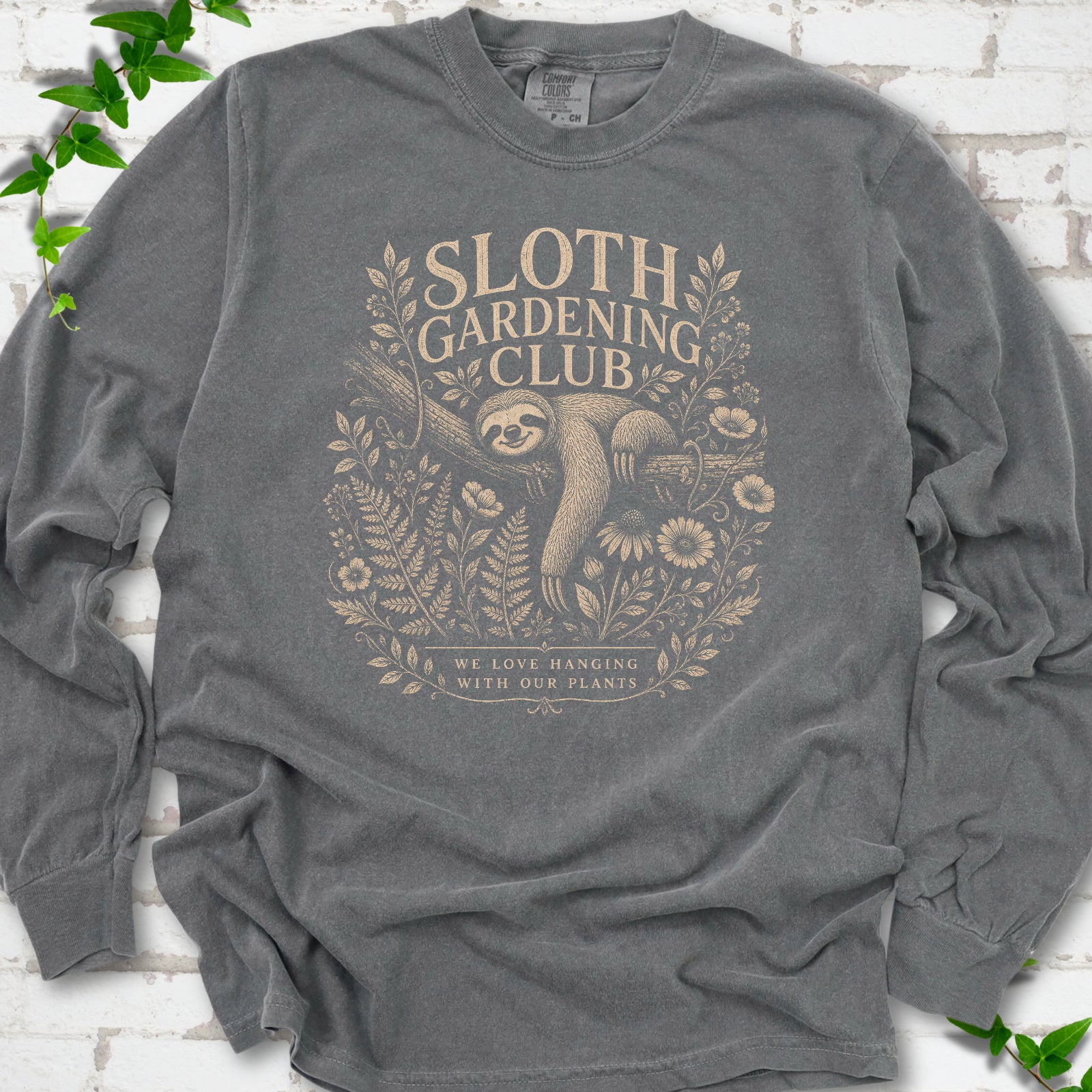 Pepper Comfort Colors® long sleeve T-shirt front mockup featuring a detailed cream-colored engraving-style illustration of a smiling sloth resting on a tree branch, surrounded by wild flowers and ferns.