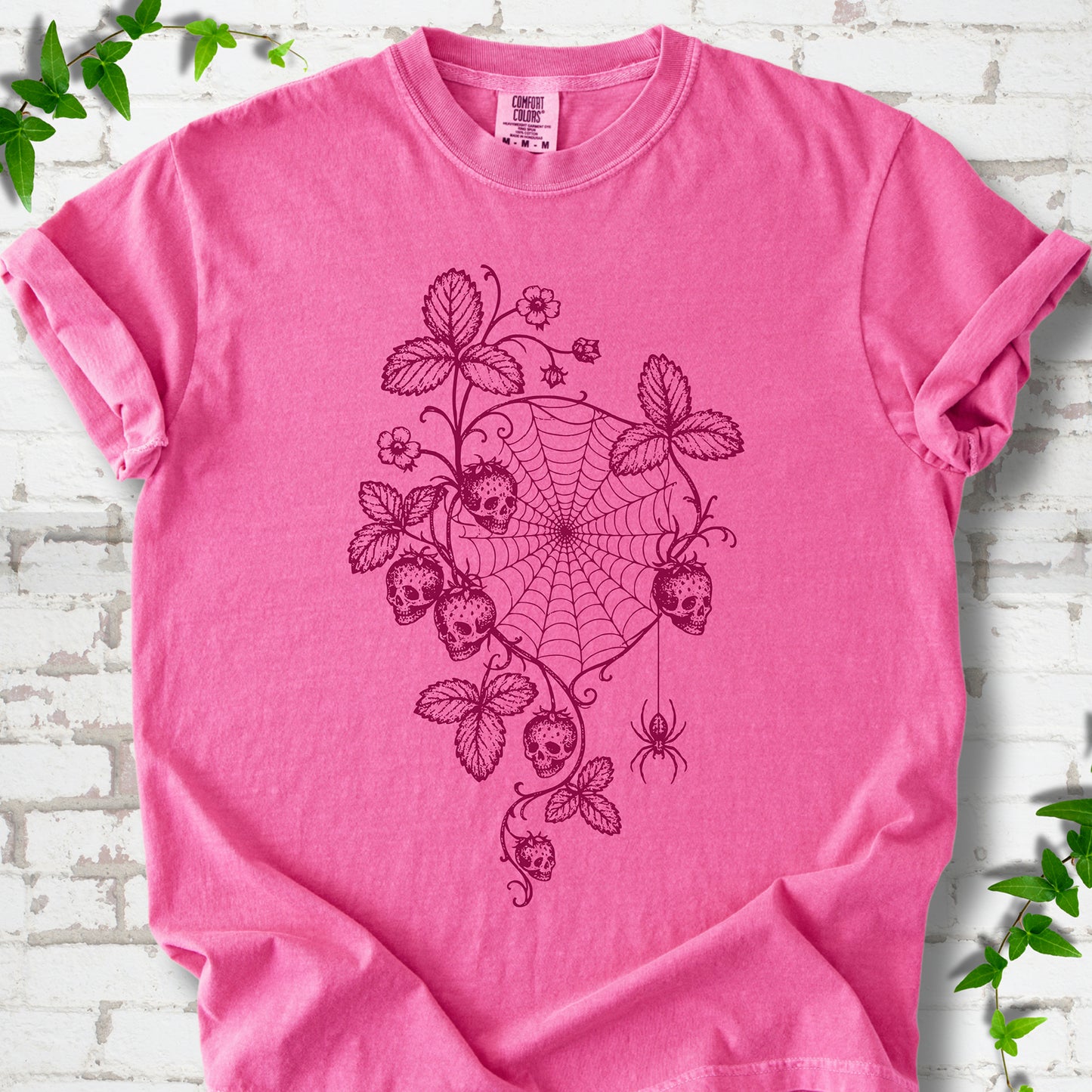 Crunchberry Comfort Colors® T-shirt flat lay featuring skull-shaped strawberries, curling leaves, a spiderweb and a hanging spider.