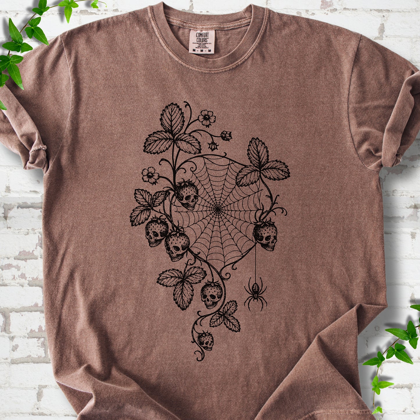 Espresso Comfort Colors® T-shirt flat lay featuring skull-shaped strawberries, curling leaves, a spiderweb and a hanging spider.