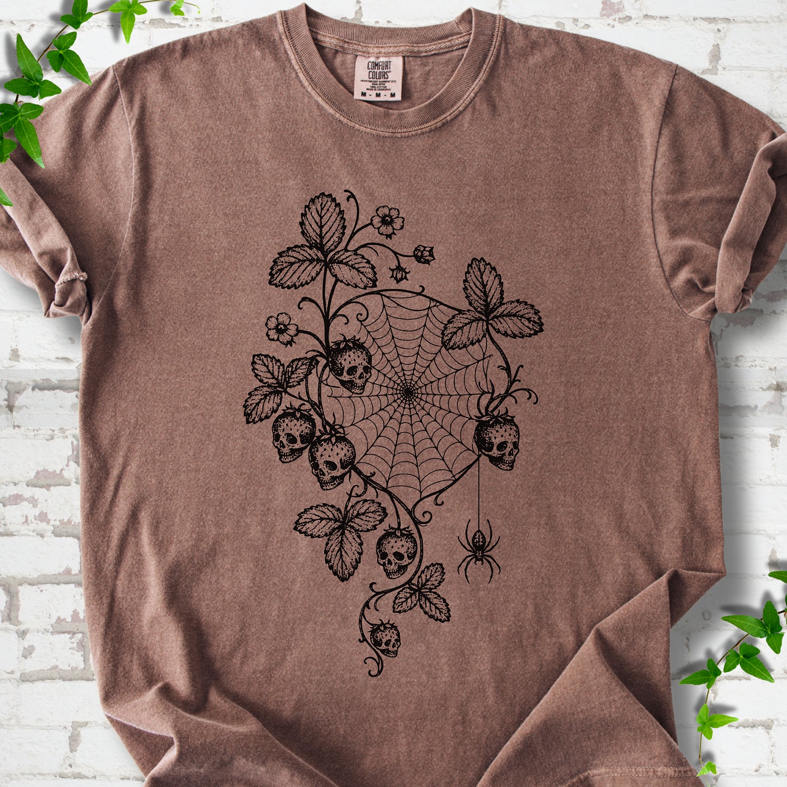 Espresso Comfort Colors® T-shirt flat lay featuring skull-shaped strawberries, curling leaves, a spiderweb and a hanging spider.