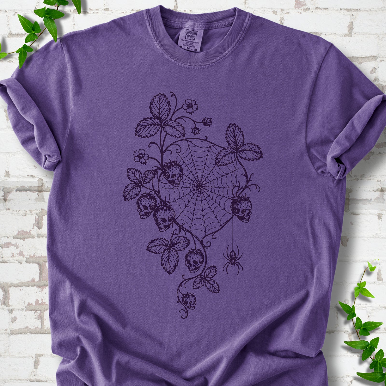 Grape Comfort Colors® T-shirt flat lay featuring skull-shaped strawberries, curling leaves, a spiderweb and a hanging spider.
