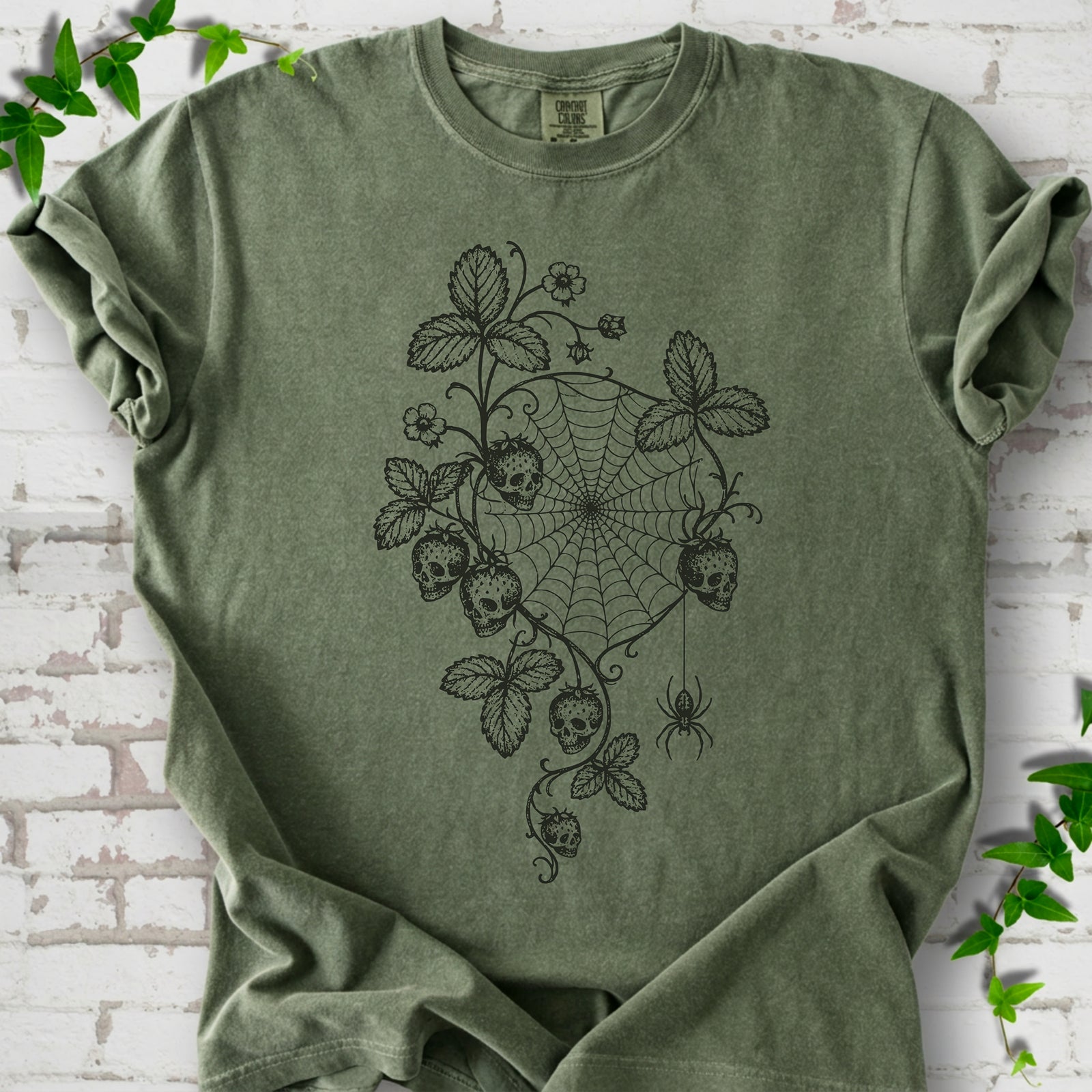 Moss Comfort Colors® T-shirt flat lay featuring skull-shaped strawberries, curling leaves, a spiderweb and a hanging spider.