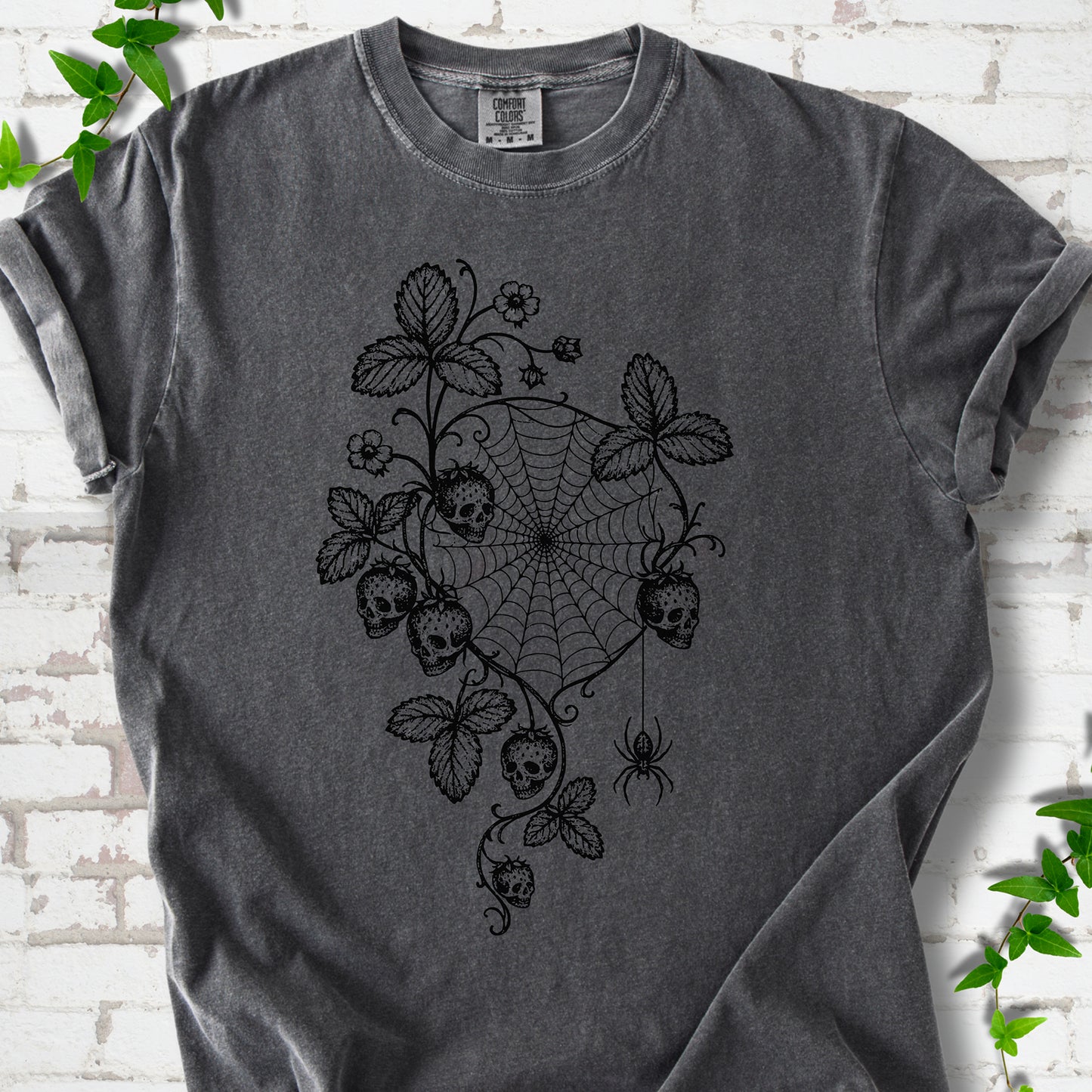 Pepper Comfort Colors® T-shirt flat lay featuring skull-shaped strawberries, curling leaves, a spiderweb and a hanging spider.