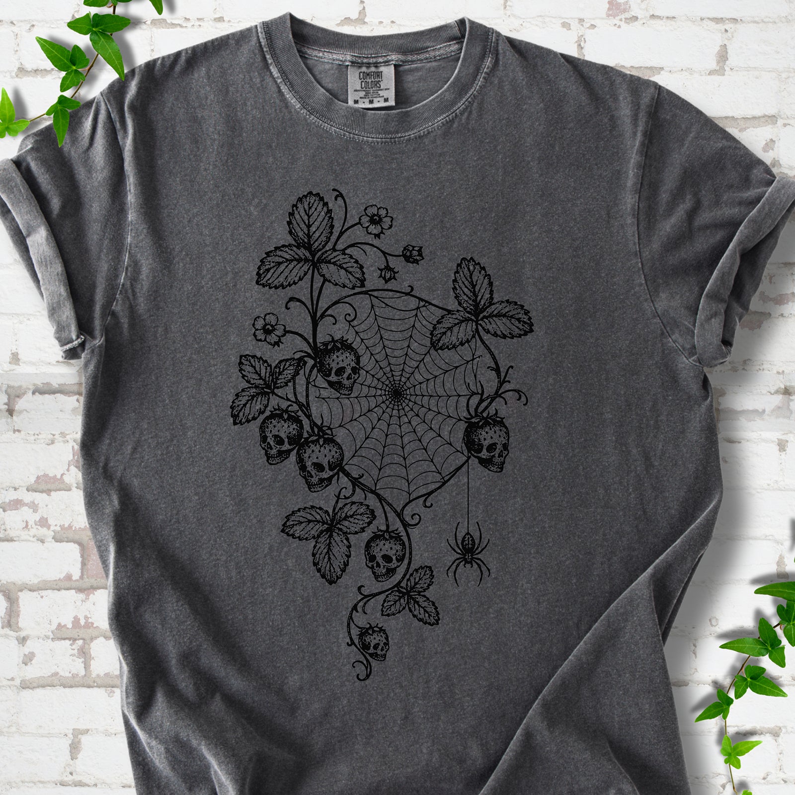 Pepper Comfort Colors® T-shirt flat lay featuring skull-shaped strawberries, curling leaves, a spiderweb and a hanging spider.