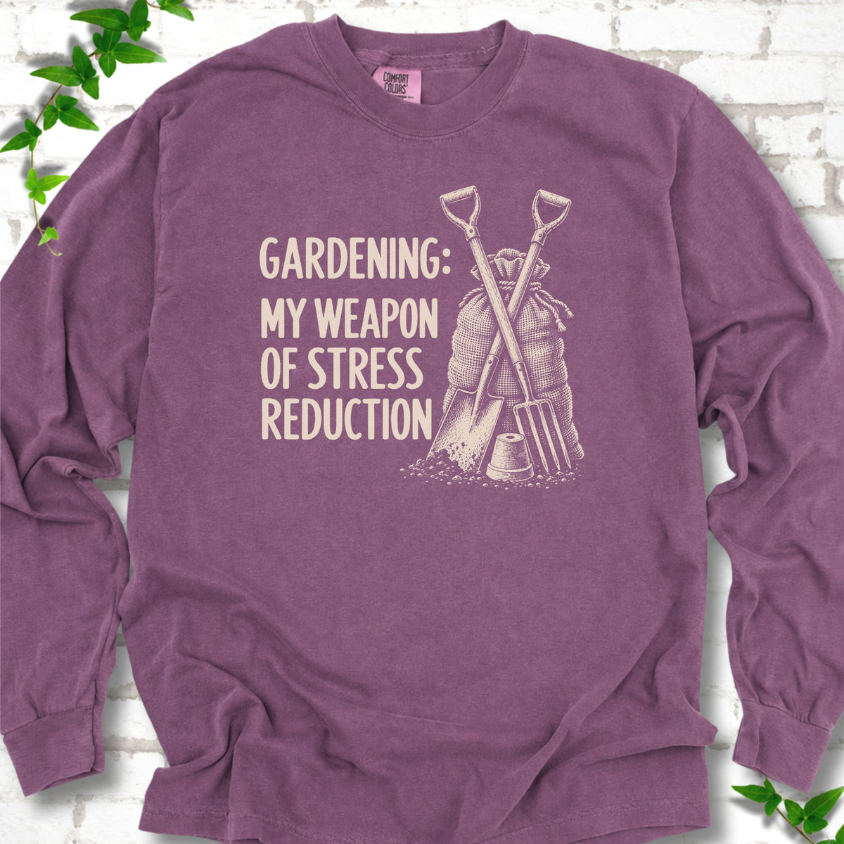 Berry long sleeve T-shirt flat lay with cream "Gardening: My Weapon of Stress Reduction" lettering beside a crossed spade and fork, burlap sack and a small flowerpot, edged by green leaves.