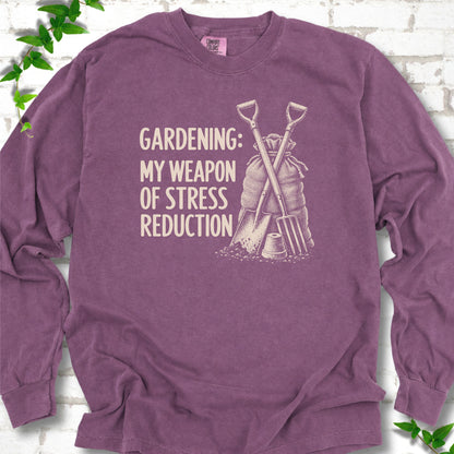 Berry long sleeve T-shirt flat lay with cream "Gardening: My Weapon of Stress Reduction" lettering beside a crossed spade and fork, burlap sack and a small flowerpot, edged by green leaves.
