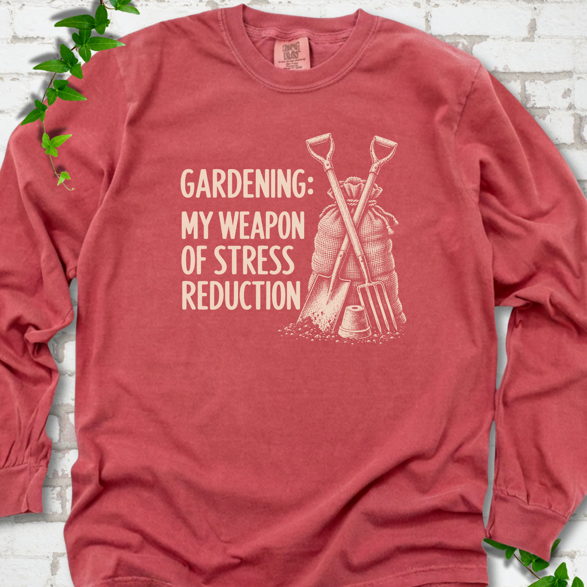 Crimson long sleeve T-shirt flat lay with cream "Gardening: My Weapon of Stress Reduction" lettering beside a crossed spade and fork, burlap sack and a small flowerpot, edged by green leaves.