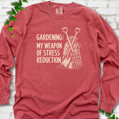 Crimson long sleeve T-shirt flat lay with cream "Gardening: My Weapon of Stress Reduction" lettering beside a crossed spade and fork, burlap sack and a small flowerpot, edged by green leaves.