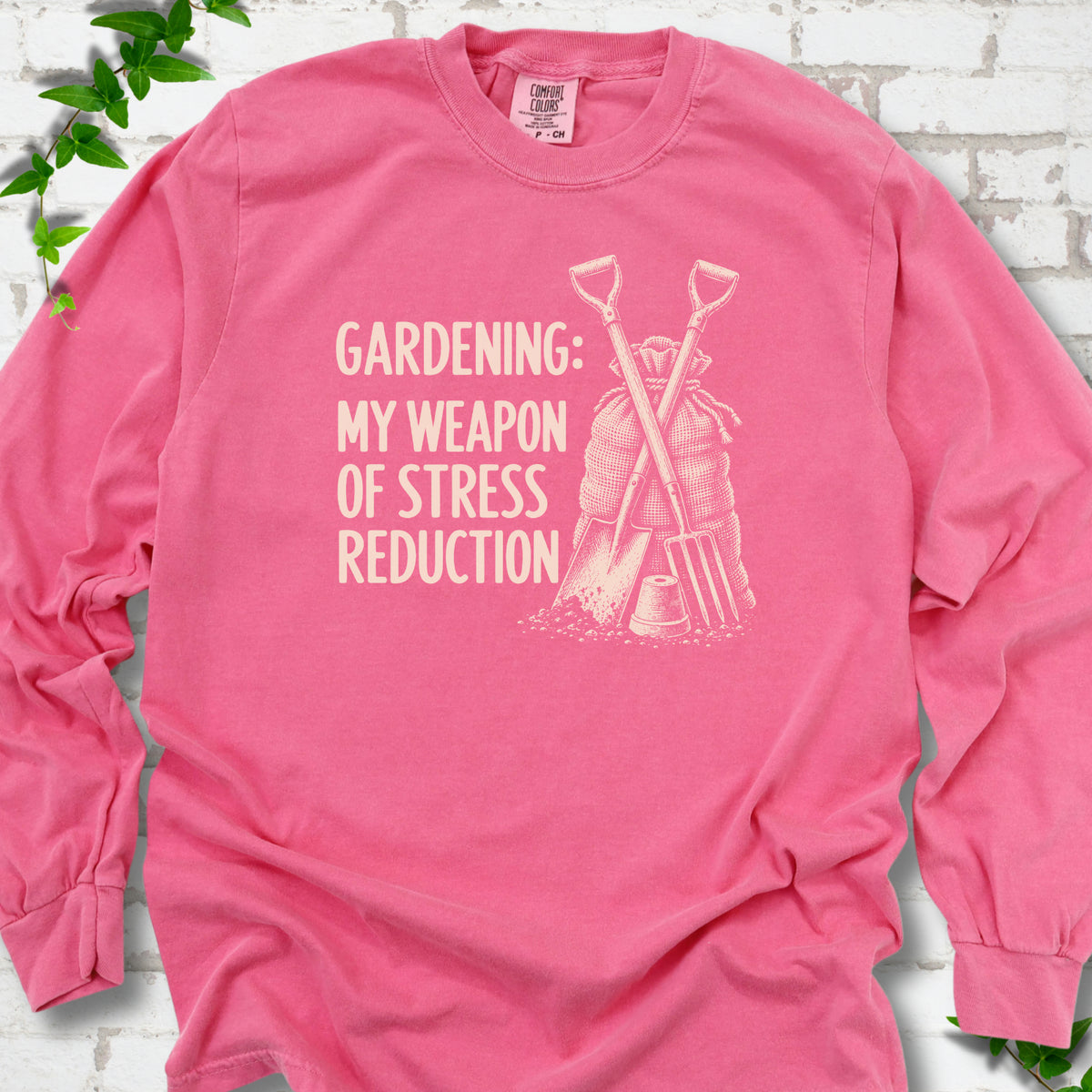 Crunchberry long sleeve T-shirt flat lay with cream "Gardening: My Weapon of Stress Reduction" lettering beside a crossed spade and fork, burlap sack and a small flowerpot, edged by green leaves.