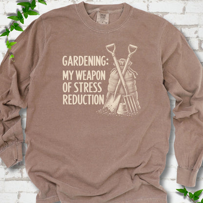 Espresso long sleeve T-shirt flat lay with cream "Gardening: My Weapon of Stress Reduction" lettering beside a crossed spade and fork, burlap sack and a small flowerpot, edged by green leaves.