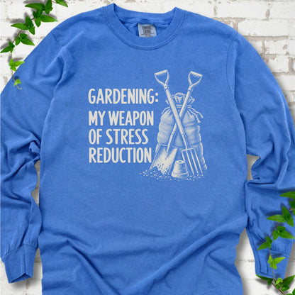 Flo Blue long sleeve T-shirt flat lay with cream "Gardening: My Weapon of Stress Reduction" lettering beside a crossed spade and fork, burlap sack and a small flowerpot, edged by green leaves.