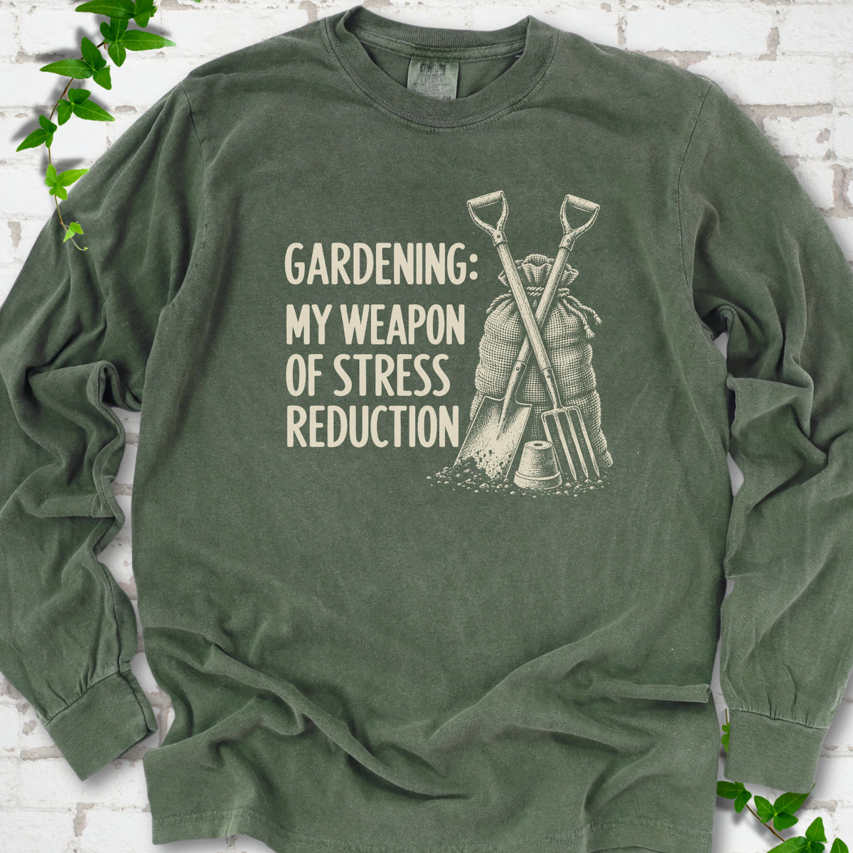 Hemp long sleeve T-shirt flat lay with cream "Gardening: My Weapon of Stress Reduction" lettering beside a crossed spade and fork, burlap sack and a small flowerpot, edged by green leaves.