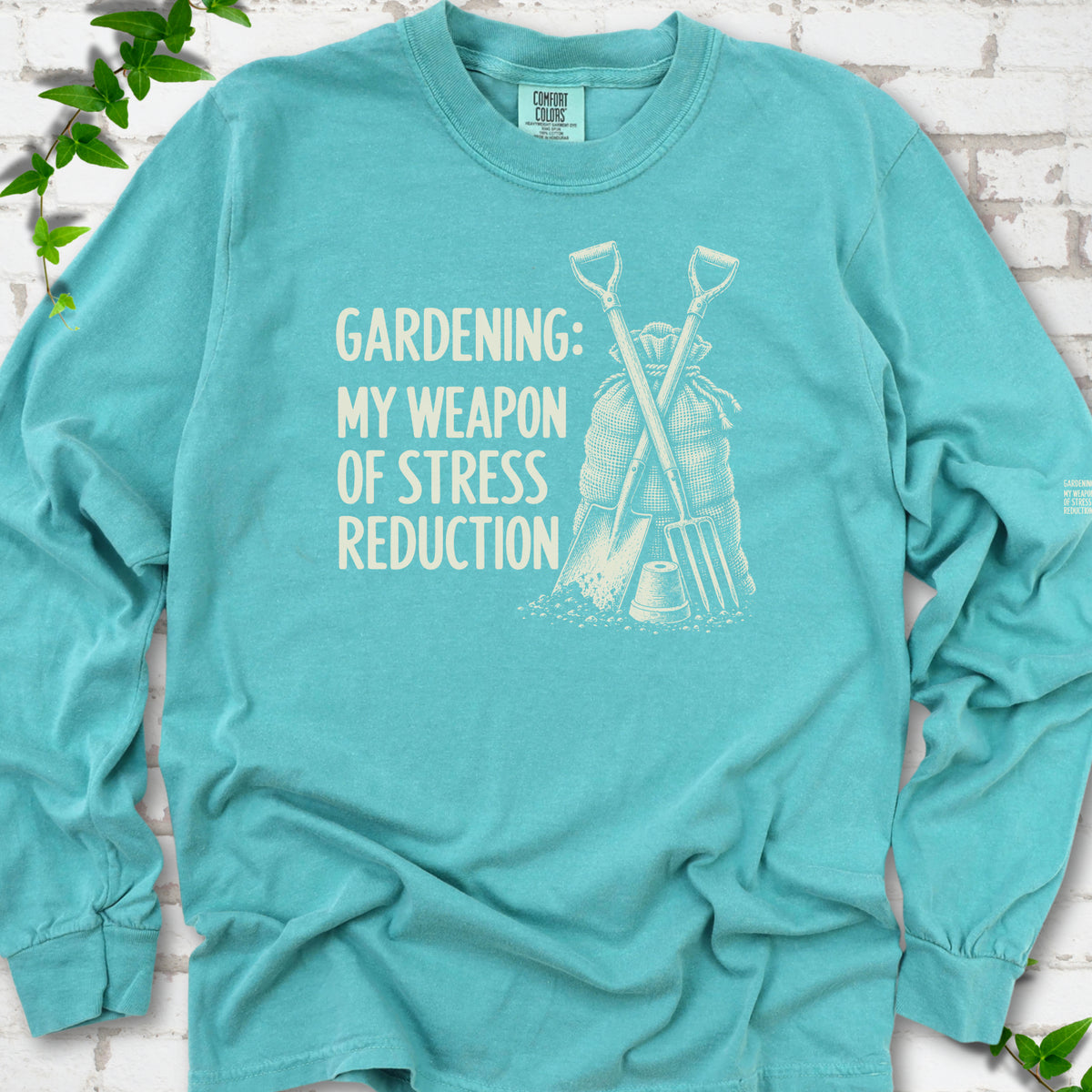 Seafoam long sleeve T-shirt flat lay with cream "Gardening: My Weapon of Stress Reduction" lettering beside a crossed spade and fork, burlap sack and a small flowerpot, edged by green leaves.