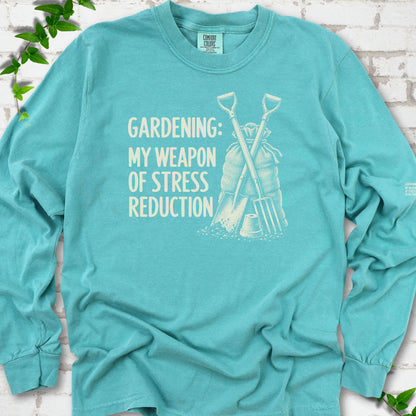 Seafoam long sleeve T-shirt flat lay with cream "Gardening: My Weapon of Stress Reduction" lettering beside a crossed spade and fork, burlap sack and a small flowerpot, edged by green leaves.