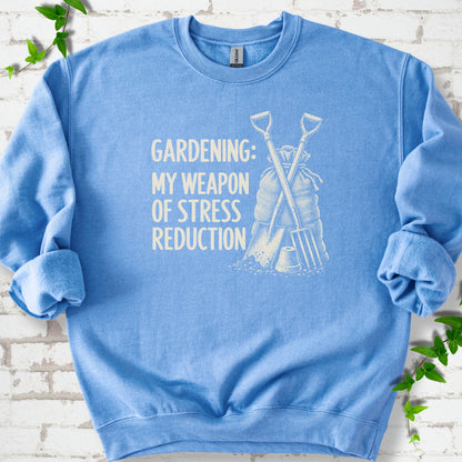 Carolina Blue sweatshirt flat lay with cream "Gardening: My Weapon of Stress Reduction" lettering beside a crossed spade and fork, burlap sack and a small flowerpot, edged by green leaves.