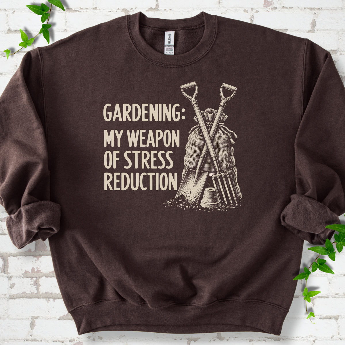 Dark Chocolate sweatshirt flat lay with cream "Gardening: My Weapon of Stress Reduction" lettering beside a crossed spade and fork, burlap sack and a small flowerpot, edged by green leaves.