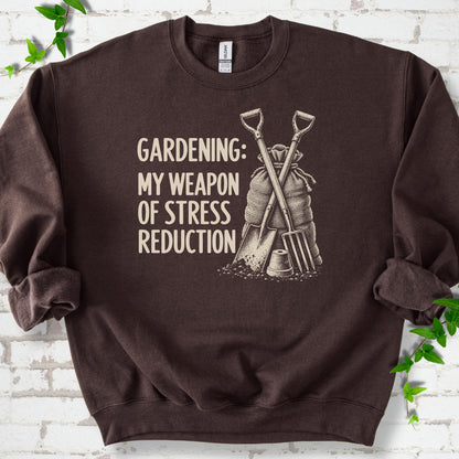 Dark Chocolate sweatshirt flat lay with cream "Gardening: My Weapon of Stress Reduction" lettering beside a crossed spade and fork, burlap sack and a small flowerpot, edged by green leaves.