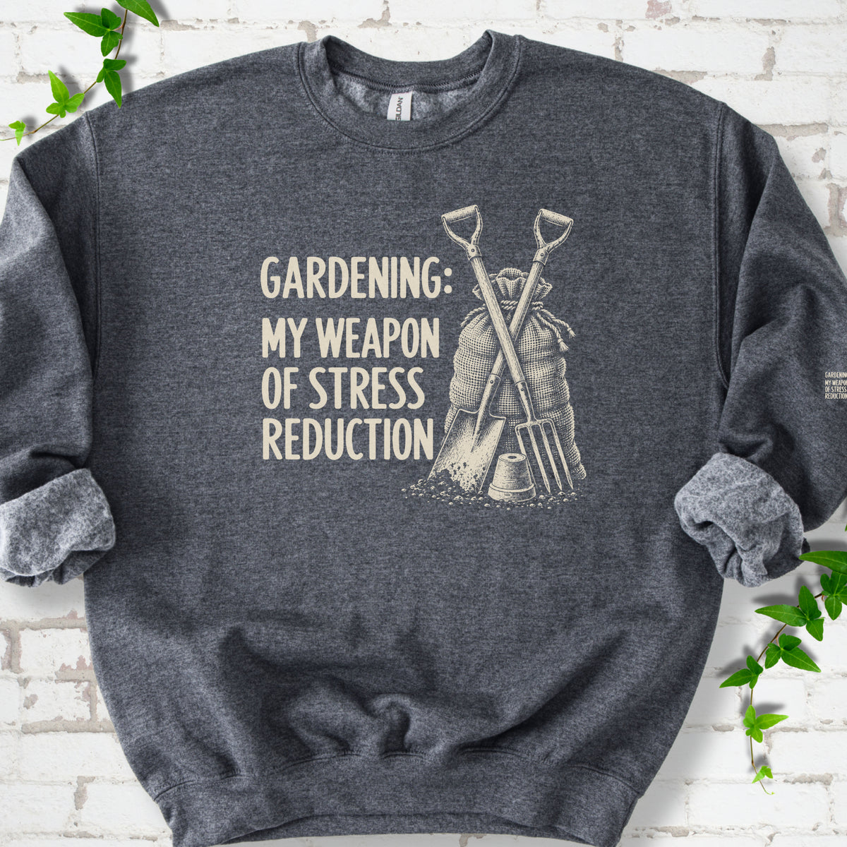 Dark Heather sweatshirt flat lay with cream "Gardening: My Weapon of Stress Reduction" lettering beside a crossed spade and fork, burlap sack and a small flowerpot, edged by green leaves.
