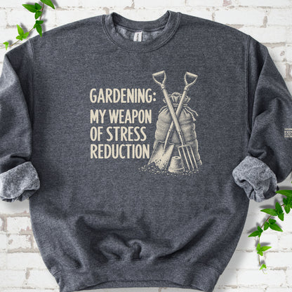 Dark Heather sweatshirt flat lay with cream "Gardening: My Weapon of Stress Reduction" lettering beside a crossed spade and fork, burlap sack and a small flowerpot, edged by green leaves.