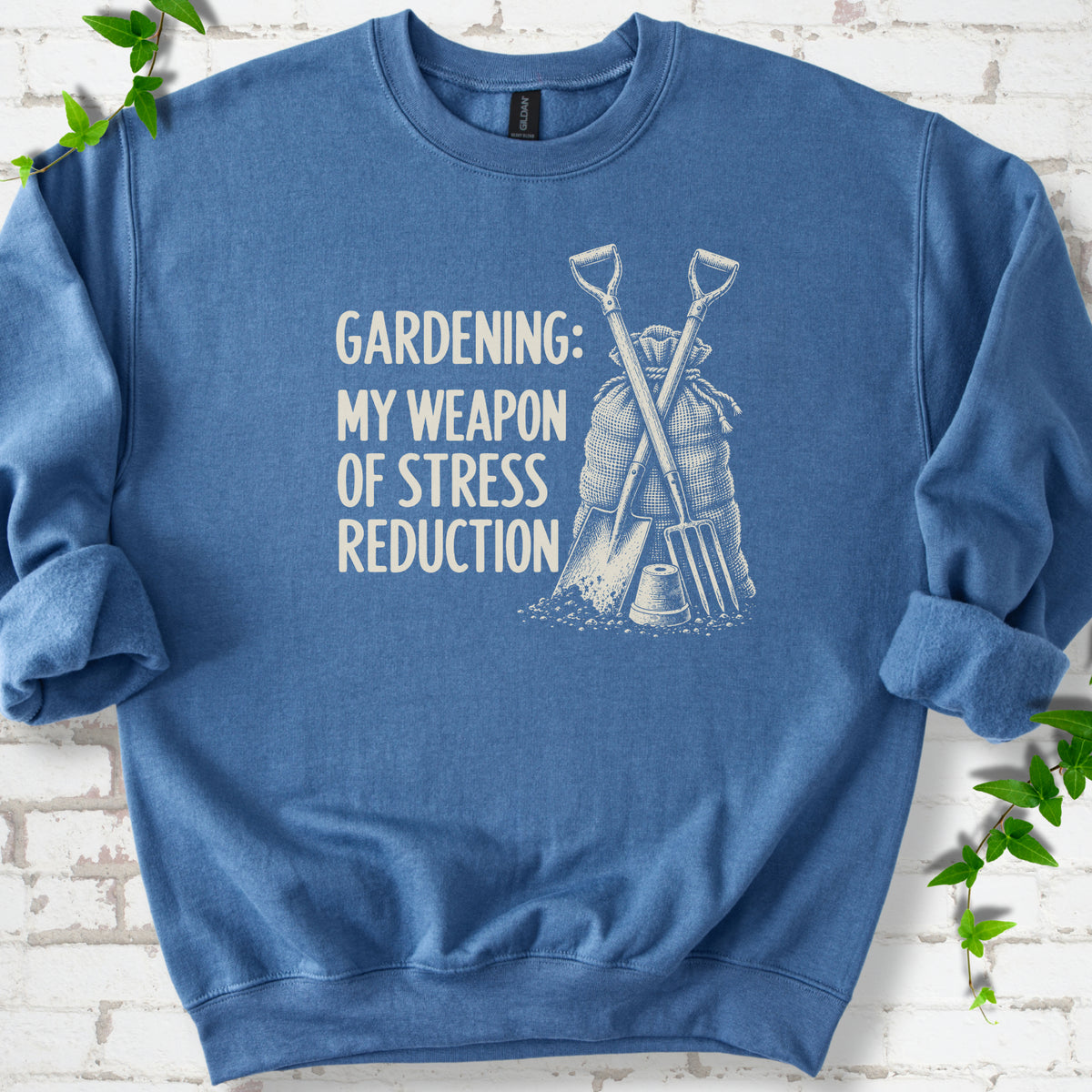 Indigo Blue sweatshirt flat lay with cream "Gardening: My Weapon of Stress Reduction" lettering beside a crossed spade and fork, burlap sack and a small flowerpot, edged by green leaves.
